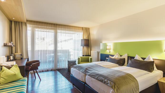 A modern hotel room with two beds, a desk, and comfortable seating. Large windows provide plenty of daylight and a view of a balcony.