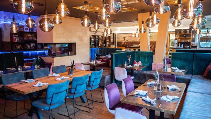 A modern restaurant with stylish tables and colorful chairs. The atmosphere is enhanced by pleasant lighting and decorative lamps.
