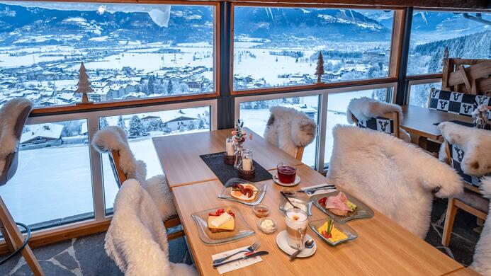 Ein gem&uuml;tlicher Tisch mit einem leckeren Fr&uuml;hst&uuml;ck in einem Bergrestaurant. Durch die gro&szlig;en Fenster hat man einen herrlichen Blick auf die schneebedeckte Landschaft.