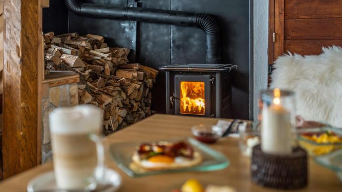 Ein gem&uuml;tlicher Raum mit einem Holzofen, der Feuer zeigt. Auf dem Tisch liegen Fr&uuml;hst&uuml;cksteller mit Lebensmitteln und eine Tasse Kaffee.