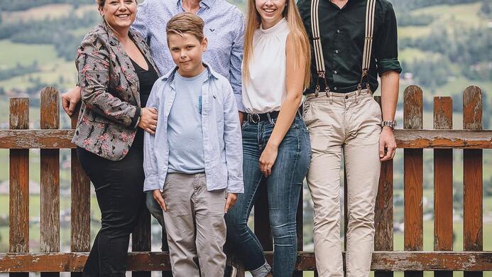 Eine Familie posiert l&auml;chelnd vor einem Holzzaun mit einer gr&uuml;nen Landschaft im Hintergrund. Ein kleiner Hund sitzt vorne bei ihnen.