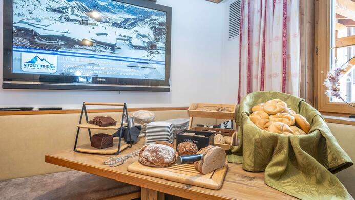 Ein reichhaltiges Fr&uuml;hst&uuml;cksbuffet mit frisch gebackenem Brot und Br&ouml;tchen. Im Hintergrund ist ein Fernseher mit einer Landschaftsaufnahme zu sehen.