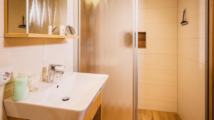 Ein modernes Badezimmer mit einer Duschkabine und einem Waschbecken. Die W&auml;nde sind hell und der Boden hat einen Holzlook.
