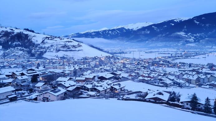 Eine verschneite Landschaft mit einem Dorf, das in sanften H&uuml;geln eingebettet ist. Der Himmel ist blau, und die Berge im Hintergrund sind mit Schnee bedeckt.