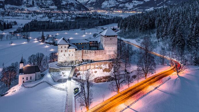Eine malerische Winterlandschaft mit einem historischen Schloss in der Mitte. Schneebedeckte Felder und B&auml;ume umgeben die Burg, w&auml;hrend eine beleuchtete Stra&szlig;e im Vordergrund verl&auml;uft.