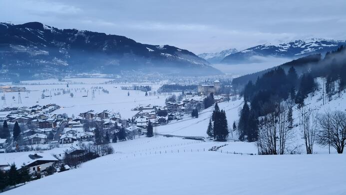 Eine verschneite Landschaft mit einem kleinen Dorf im Tal. Umgeben von Bergen und einem nebligen Himmel.