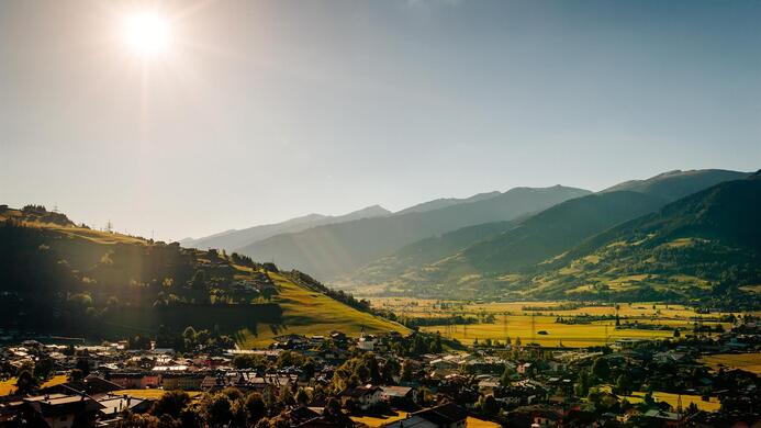 Eine malerische Landschaft mit H&uuml;geln und Bergen im Hintergrund. Die Sonne scheint &uuml;ber ein ruhiges, gr&uuml;nes Tal und ein kleines Dorf.