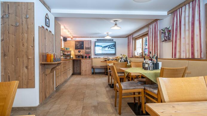 Ein gem&uuml;tliches Restaurant mit Holzdekor und einfachen Tischen. Im Hintergrund ist eine Theke und ein Fernseher, der eine Berglandschaft zeigt.