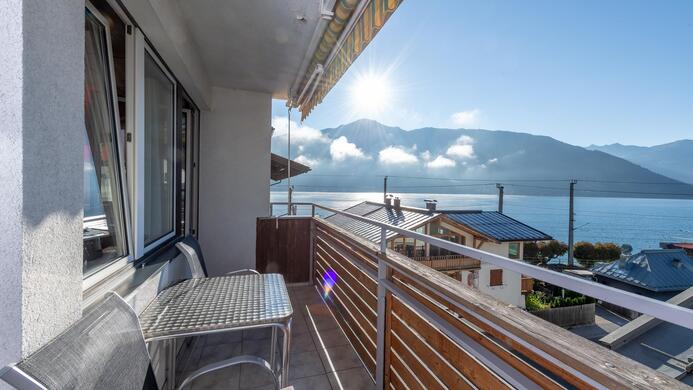 Ein Balkon mit einem Tisch und St&uuml;hlen bietet einen Blick auf einen klaren See und Berge im Hintergrund. Der Himmel ist sonnig und blau.