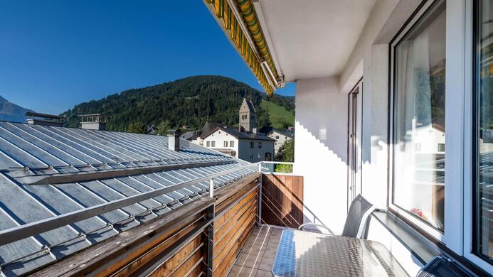 Ein Balkon mit einem Blick auf Berge und ein Dorf. Die Sonne scheint und der Himmel ist klar.