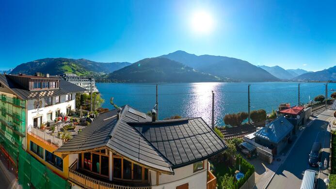 Eine malerische Seelandschaft mit Bergen im Hintergrund. In der Sonne reflektiert das Wasser, w&auml;hrend die H&auml;user am Ufer stehen.