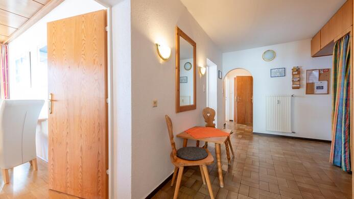 Ein heller Flur mit einem Holzfu&szlig;boden und einer kleinen Sitzecke. An der Wand h&auml;ngen ein Spiegel und einige Informationsbl&auml;tter.