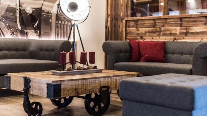 A modernly furnished living space with gray sofas and a wooden table on wheels. On the table are candles, and in the background, an elegant lamp can be seen.